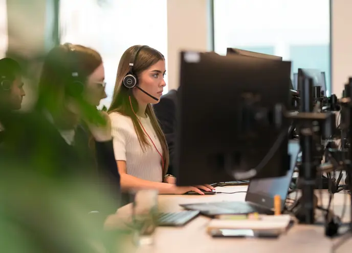 A woman wearing headset and sitting at a desk with other people in a modern office.